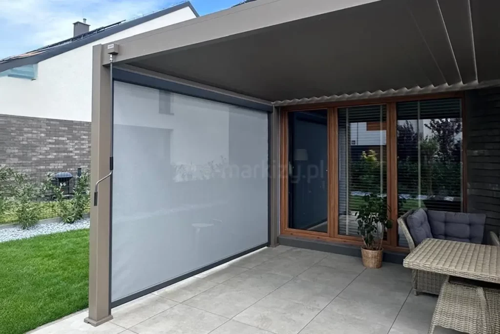 Nowoczesna pergola przyścienna Marlo zamontowana na tarasie wyłożonym jasnoszarymi płytami gładkimi. Konstrukcja pergoli wykonana jest z profili aluminiowych w kolorze beżowym lub jasnobrązowym, a jej zadaszenie stanowi ruchomy, segmentowy dach tkaninowy w ciemniejszym odcieniu brązu. Na lewym boku konstrukcji zainstalowano pionową roletę typu screen w kolorze jasnoszarym, która jest częściowo transparentna i pełni funkcję zabudowy bocznej chroniącej przed słońcem i wiatrem. Do obsługi rolety służy widoczna po lewej stronie korba ręczna. Pod zadaszeniem znajduje się strefa wypoczynkowa z technorattanowymi meblami ogrodowymi w kolorze szarym, w tym sofa z poduszkami oraz stolik. W tle widoczna jest elewacja domu z dużymi przeszkleniami w brązowych ramach, fragment trawnika oraz ogrodzenie z antracytowej cegły.