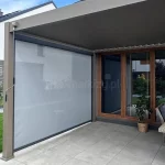 Nowoczesna pergola przyścienna Marlo zamontowana na tarasie wyłożonym jasnoszarymi płytami gładkimi. Konstrukcja pergoli wykonana jest z profili aluminiowych w kolorze beżowym lub jasnobrązowym, a jej zadaszenie stanowi ruchomy, segmentowy dach tkaninowy w ciemniejszym odcieniu brązu. Na lewym boku konstrukcji zainstalowano pionową roletę typu screen w kolorze jasnoszarym, która jest częściowo transparentna i pełni funkcję zabudowy bocznej chroniącej przed słońcem i wiatrem. Do obsługi rolety służy widoczna po lewej stronie korba ręczna. Pod zadaszeniem znajduje się strefa wypoczynkowa z technorattanowymi meblami ogrodowymi w kolorze szarym, w tym sofa z poduszkami oraz stolik. W tle widoczna jest elewacja domu z dużymi przeszkleniami w brązowych ramach, fragment trawnika oraz ogrodzenie z antracytowej cegły.