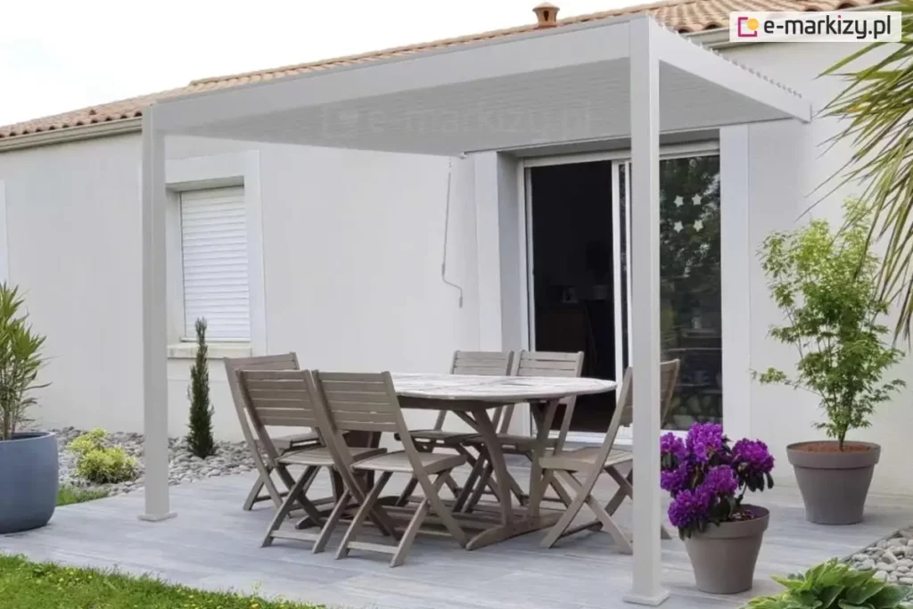 Biała pergola przyścienna zamontowana na nowoczesnym tarasie wyłożonym jasnoszarymi deskami. Konstrukcja wspiera się na dwóch pionowych słupach od strony ogrodu i jest przymocowana bezpośrednio do białej elewacji budynku nad drzwiami przesuwnymi. Pod zadaszeniem znajduje się drewniany zestaw mebli ogrodowych: duży owalny stół oraz sześć krzeseł z listewkami. Na tarasie ustawiono dekoracyjne rośliny w donicach, w tym intensywnie fioletowe kwiaty oraz zielone drzewka ozdobne. W tle widoczna jest biała ściana domu z oknem wyposażonym w białą roletę zewnętrzną oraz fragment dachu z dachówki ceramicznej. Z górnej belki pergoli zwisa biały drążek korbowy służący do regulacji kąta nachylenia lamel dachu.