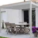 Biała pergola przyścienna zamontowana na nowoczesnym tarasie wyłożonym jasnoszarymi deskami. Konstrukcja wspiera się na dwóch pionowych słupach od strony ogrodu i jest przymocowana bezpośrednio do białej elewacji budynku nad drzwiami przesuwnymi. Pod zadaszeniem znajduje się drewniany zestaw mebli ogrodowych: duży owalny stół oraz sześć krzeseł z listewkami. Na tarasie ustawiono dekoracyjne rośliny w donicach, w tym intensywnie fioletowe kwiaty oraz zielone drzewka ozdobne. W tle widoczna jest biała ściana domu z oknem wyposażonym w białą roletę zewnętrzną oraz fragment dachu z dachówki ceramicznej. Z górnej belki pergoli zwisa biały drążek korbowy służący do regulacji kąta nachylenia lamel dachu.