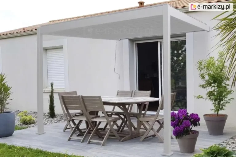 Biała pergola przyścienna zamontowana na nowoczesnym tarasie wyłożonym jasnoszarymi deskami. Konstrukcja wspiera się na dwóch pionowych słupach od strony ogrodu i jest przymocowana bezpośrednio do białej elewacji budynku nad drzwiami przesuwnymi. Pod zadaszeniem znajduje się drewniany zestaw mebli ogrodowych: duży owalny stół oraz sześć krzeseł z listewkami. Na tarasie ustawiono dekoracyjne rośliny w donicach, w tym intensywnie fioletowe kwiaty oraz zielone drzewka ozdobne. W tle widoczna jest biała ściana domu z oknem wyposażonym w białą roletę zewnętrzną oraz fragment dachu z dachówki ceramicznej. Z górnej belki pergoli zwisa biały drążek korbowy służący do regulacji kąta nachylenia lamel dachu.