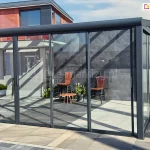 konstrukcja nowoczesnego zadaszenia ogrodowego wyposażona w system szklanych drzwi przesuwnych Sotro na frontowej ścianie. Aluminiowa rama w kolorze grafitowym tworzy stabilny szkielet dla dużych, transparentnych tafli szkła, które odbijają geometryczne cienie rzucane przez konstrukcję dachu. Wewnątrz przeszklonej altany widoczna jest minimalistyczna aranżacja z dwoma drewnianymi krzesłami o wysokich oparciach i niskim stolikiem kawowym, ustawionymi na posadzce z dużych, jasnych płyt tarasowych. Boczna ściana wykonana jest z ciemnych paneli o strukturze kamienia, co nadaje całości luksusowy charakter. Szerokie skrzydła przesuwne zamontowane na wielotorowej szynie dolnej pozwalają na swobodną regulację przepływu powietrza i łatwy dostęp do wnętrza, tworząc funkcjonalny salon ogrodowy.