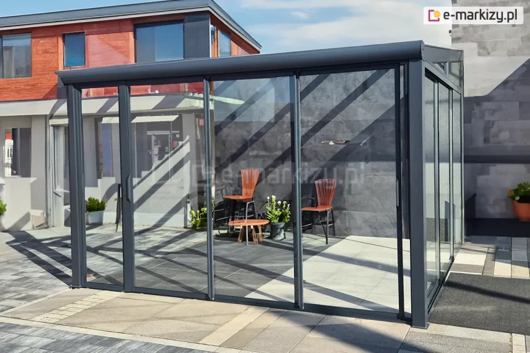 konstrukcja nowoczesnego zadaszenia ogrodowego wyposażona w system szklanych drzwi przesuwnych Sotro na frontowej ścianie. Aluminiowa rama w kolorze grafitowym tworzy stabilny szkielet dla dużych, transparentnych tafli szkła, które odbijają geometryczne cienie rzucane przez konstrukcję dachu. Wewnątrz przeszklonej altany widoczna jest minimalistyczna aranżacja z dwoma drewnianymi krzesłami o wysokich oparciach i niskim stolikiem kawowym, ustawionymi na posadzce z dużych, jasnych płyt tarasowych. Boczna ściana wykonana jest z ciemnych paneli o strukturze kamienia, co nadaje całości luksusowy charakter. Szerokie skrzydła przesuwne zamontowane na wielotorowej szynie dolnej pozwalają na swobodną regulację przepływu powietrza i łatwy dostęp do wnętrza, tworząc funkcjonalny salon ogrodowy.