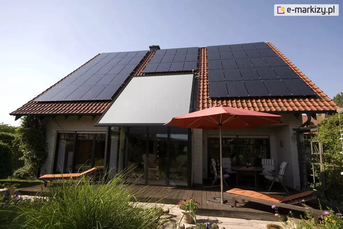 Szerokie ujęcie elewacji tylnej domu jednorodzinnego z dachem dwuspadowym pokrytym czerwoną dachówką ceramiczną, na którym zainstalowano rozbudowany system paneli fotowoltaicznych oraz nowoczesną roletę veranda V-Ziiip. Veranda z tkaniną w kolorze jasnoszarym jest całkowicie rozwinięta i zamontowana bezpośrednio nad przeszklonym wykuszem lub ogrodem zimowym o ciemnych profilach, stanowiąc integralną część połaci dachowej. Po obu stronach markizy znajdują się liczne rzędy ciemnych modułów fotowoltaicznych, które wypełniają niemal całą dostępną powierzchnię dachu. Przed budynkiem rozciąga się przestronny taras wykonany z ciemnego drewna, na którym ustawiono meble ogrodowe, stół oraz leżaki z pomarańczowymi materacami. Czerwony parasol ogrodowy rozłożony nad stołem koresponduje kolorystycznie z barwą dachówki. Teren wokół tarasu jest bogato obsadzony zielenią, w tym wysokimi trawami ozdobnymi i krzewami, co nadaje posesji przytulny charakter. Całość sceny jest oświetlona jasnym, popołudniowym światłem przy bezchmurnym, błękitnym niebie, co podkreśla nowoczesną architekturę i proekologiczne rozwiązania zastosowane w budynku.