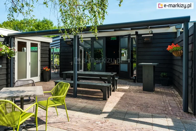 Zdjęcie ukazuje zewnętrzną strefę wypoczynkową przylegającą do budynku o elewacji z poziomych, czarnych desek, osłoniętą nowoczesnym zadaszeniem tarasowym Nebbiolo marki Deponti. Konstrukcja zadaszenia wykonana jest z ciemnoszarych profili aluminiowych o matowym wykończeniu, z charakterystyczną zaokrągloną rynną frontową podtrzymywaną przez pionowe, smukłe słupy. Przestronny taras wyłożony postarzaną kostką brukową został wyposażony w czarny, drewniany stół z dwiema ławami oraz nowoczesne krzesła w kolorze intensywnej limonki i czerni ustawione przy mniejszych stolikach. Na czarnej ścianie budynku zamontowano promienniki ciepła oraz zawieszono donice z kwitnącymi, czerwonymi kwiatami, które kontrastują z ciemną elewacją. W tle, za otwartymi przeszklonymi drzwiami prowadzącymi do wnętrza, widoczna jest gęsta zieleń ogrodowa oraz gałęzie brzozy zwisające nad konstrukcją zadaszenia w pełnym świetle dziennym.