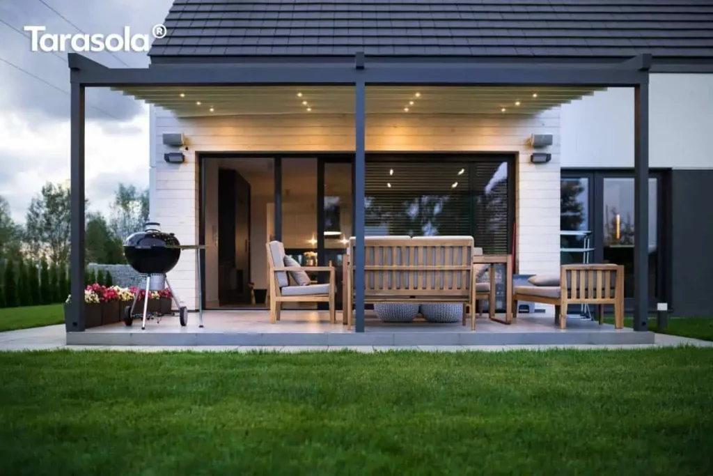 Widok od strony ogrodu na nowoczesną, dwumodułową pergolę przyścienną Elegancy Tarasola, zamontowaną przy budynku o jasnej elewacji wyłożonej poziomymi deskami. Konstrukcja z ekstrudowanego aluminium w kolorze antracytowym posiada zadaszenie z jasnej tkaniny, pod którą rozmieszczono zintegrowane punkty świetlne LED emitujące ciepły blask. Na betonowym tarasie znajduje się strefa wypoczynkowa z drewnianymi sofami o szarych siedziskach oraz czarny grill kulisty ustawiony po lewej stronie. Przeszklone, duże drzwi przesuwne prowadzą do wnętrza domu, a przed tarasem rozciąga się równo przystrzyżony trawnik. Całość kompozycji domyka pas niskich donic z różowymi i białymi kwiatami oraz ciemny, dwuspadowy dach budynku.
