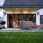 Widok od strony ogrodu na nowoczesną, dwumodułową pergolę przyścienną Elegancy Tarasola, zamontowaną przy budynku o jasnej elewacji wyłożonej poziomymi deskami. Konstrukcja z ekstrudowanego aluminium w kolorze antracytowym posiada zadaszenie z jasnej tkaniny, pod którą rozmieszczono zintegrowane punkty świetlne LED emitujące ciepły blask. Na betonowym tarasie znajduje się strefa wypoczynkowa z drewnianymi sofami o szarych siedziskach oraz czarny grill kulisty ustawiony po lewej stronie. Przeszklone, duże drzwi przesuwne prowadzą do wnętrza domu, a przed tarasem rozciąga się równo przystrzyżony trawnik. Całość kompozycji domyka pas niskich donic z różowymi i białymi kwiatami oraz ciemny, dwuspadowy dach budynku.