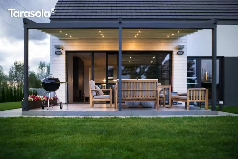 Widok od strony ogrodu na nowoczesną, dwumodułową pergolę przyścienną Elegancy Tarasola, zamontowaną przy budynku o jasnej elewacji wyłożonej poziomymi deskami. Konstrukcja z ekstrudowanego aluminium w kolorze antracytowym posiada zadaszenie z jasnej tkaniny, pod którą rozmieszczono zintegrowane punkty świetlne LED emitujące ciepły blask. Na betonowym tarasie znajduje się strefa wypoczynkowa z drewnianymi sofami o szarych siedziskach oraz czarny grill kulisty ustawiony po lewej stronie. Przeszklone, duże drzwi przesuwne prowadzą do wnętrza domu, a przed tarasem rozciąga się równo przystrzyżony trawnik. Całość kompozycji domyka pas niskich donic z różowymi i białymi kwiatami oraz ciemny, dwuspadowy dach budynku.