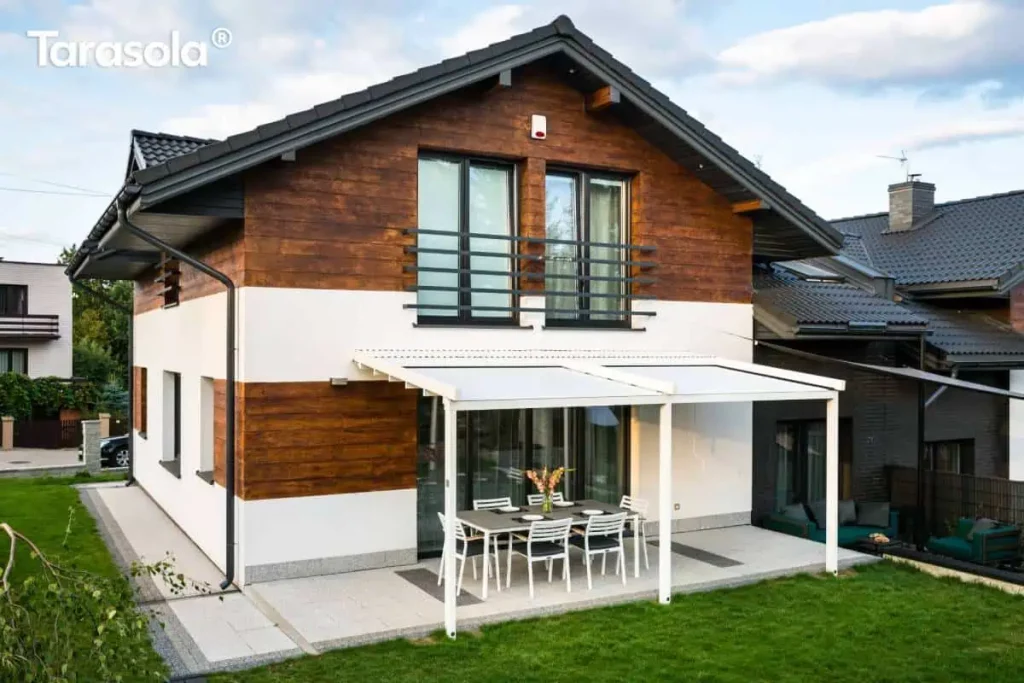 Nowoczesna przyścienna pergola tarasowa Tarasola Simple w kolorze białym zamontowana na elewacji piętrowego domu jednorodzinnego. Konstrukcja wspiera się na dwóch pionowych słupach o kwadratowym przekroju, a jej zadaszenie stanowi jasna tkanina techniczna rozpięta na aluminiowych profilach. Budynek posiada elewację łączącą biały tynk z poziomymi panelami o strukturze ciemnego drewna oraz dwuspadowy dach pokryty ciemną dachówką. Pod pergolą, na tarasie wyłożonym jasnoszarymi płytami z ciemniejszymi detalami, ustawiono stół jadalniany z sześcioma białymi krzesłami ogrodowymi. Szerokie przeszklenie tarasowe zapewnia bezpośrednie wyjście z wnętrza domu pod zadaszenie. Wokół tarasu rozciąga się zadbany trawnik, a w tle widoczna jest zabudowa sąsiednia oraz fragmenty zieleni ogrodowej.
