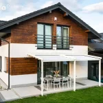 Nowoczesna przyścienna pergola tarasowa Tarasola Simple w kolorze białym zamontowana na elewacji piętrowego domu jednorodzinnego. Konstrukcja wspiera się na dwóch pionowych słupach o kwadratowym przekroju, a jej zadaszenie stanowi jasna tkanina techniczna rozpięta na aluminiowych profilach. Budynek posiada elewację łączącą biały tynk z poziomymi panelami o strukturze ciemnego drewna oraz dwuspadowy dach pokryty ciemną dachówką. Pod pergolą, na tarasie wyłożonym jasnoszarymi płytami z ciemniejszymi detalami, ustawiono stół jadalniany z sześcioma białymi krzesłami ogrodowymi. Szerokie przeszklenie tarasowe zapewnia bezpośrednie wyjście z wnętrza domu pod zadaszenie. Wokół tarasu rozciąga się zadbany trawnik, a w tle widoczna jest zabudowa sąsiednia oraz fragmenty zieleni ogrodowej.