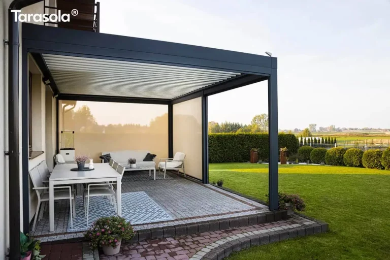 Zdjęcie przedstawia nowoczesną aranżację tarasu z wykorzystaniem zadaszenia Essential tarasola w formie wolnostojącej konstrukcji opartej o ścianę budynku. Aluminiowa rama pergoli w głębokim kolorze antracytowym kontrastuje z białymi, ruchomymi lamelami dachowymi, które są całkowicie zamknięte, tworząc szczelny dach. Konstrukcja została wyposażona w boczne przesłony w formie rolet screen w jasnobeżowym kolorze; jedna z nich jest całkowicie opuszczona, zasłaniając tylną część tarasu, natomiast boczna roleta jest podniesiona, otwierając widok na ogród. Na podwyższonym tarasie wybrukowanym jasnoszarą kostką z ciemniejszym obramowaniem wydzielono dwie strefy. W części jadalnianej, na wzorzystym niebiesko-białym dywanie, stoi duży biały stół z dopasowanymi krzesłami, na którym umieszczono dekoracyjne doniczki i świecę. W głębi, pod opuszczoną roletą, znajduje się strefa wypoczynkowa z białą sofą, fotelem i stolikiem kawowym na drugim dywanie. Po lewej stronie widoczny jest fragment elewacji domu z rynną spustową i donicami z różowymi kwiatami. Tło stanowi rozległy, zielony trawnik, niski żywopłot oraz otwarty krajobraz pól pod jasnym, pogodnym niebem.