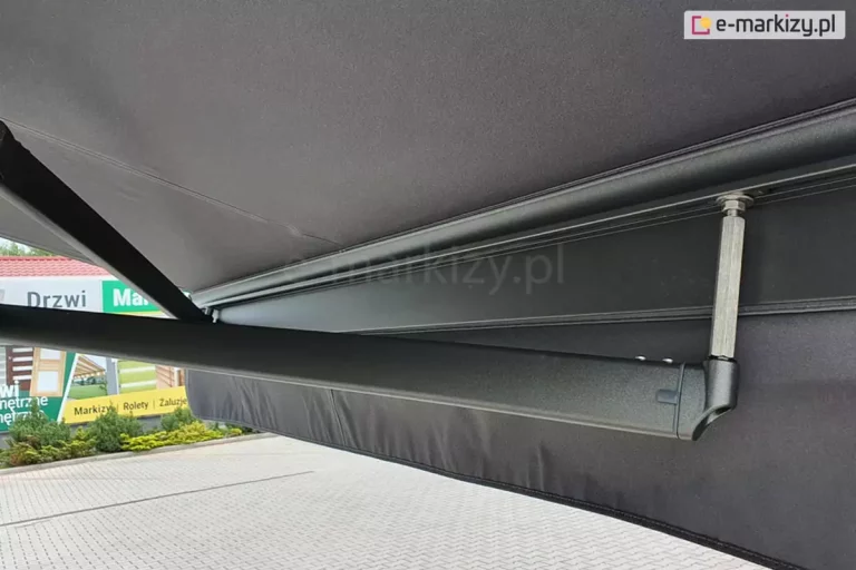 Ujęta od boku, od strony obniżonego ramienia krzyżowego belka przednia markizy Palladio Selt widoczna od wnętrza markizy w kolorze grafitowym konstrukcji i poszycia z prostą falbaną. Wyraźnie widoczne skrzyżowanie ramion oraz wydłużone mocowanie jednego z ramion w belce przedniej.