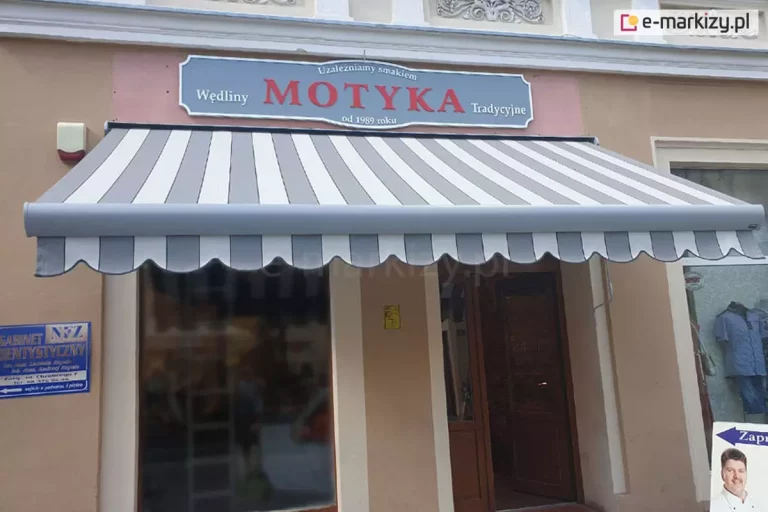 Zdjęcie prezentuje frontową elewację punktu handlowego zlokalizowanego w kamienicy, nad którym zamontowana została klasyczna markiza tarasowa w pełnym rozwinięciu. Poszycie markizy charakteryzuje się regularnym wzorem w pionowe, szerokie pasy w kolorach białym oraz jasnoszarym. Dolna krawędź tkaniny wykończona jest ozdobną, falistą falbaną, która kontynuuje pasiasty motyw i posiada ciemniejszą lamówkę na samym brzegu. Konstrukcja nośna markizy, w tym profil przedni, jest wykonana w kolorze jasnoszarym, spójnym z jednym z kolorów pasów. Powyżej markizy, na beżowej elewacji, znajduje się duży, szary szyld z czerwonym napisem „MOTYKA” oraz dodatkowymi informacjami o tradycyjnych wędlinach. Pod zadaszeniem widoczne są przeszklone drzwi wejściowe do lokalu oraz duża witryna sklepowa, w której odbijają się elementy ulicy. Po lewej stronie od wejścia na ścianie zamontowana jest niebieska tablica informacyjna gabinetu dentystycznego oraz mały alarmowy sygnalizator świetlny. Po prawej stronie widać fragment sąsiedniej witryny z męską koszulą na wystawie oraz część potykacza reklamowego ze zdjęciem uśmiechniętego mężczyzny. Całość kompozycji oddaje wygląd miejskiej ulicy handlowej w dzień.