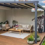 Zdjęcie prezentuje nowoczesną pergolę tarasową Aura o konstrukcji aluminiowej w kolorze ciemnego antracytu, zamontowaną do jasnej elewacji budynku. Pergola posiada zadaszenie tkaninowe w odcieniu beżowym, które jest rozpięte na poprzecznych profilach przeciwwietrznych. Charakterystycznym elementem tego modelu jest zintegrowane oświetlenie LED w formie punktowych diod umieszczonych w profilach podtrzymujących materiał, co tworzy nastrojową atmosferę po zmroku. Konstrukcja wspiera się na solidnych pionowych słupach o kwadratowym przekroju, usytuowanych przy zewnętrznej krawędzi tarasu. Po lewej stronie, przy ścianie budynku, widoczna jest długa korba służąca do manualnego sterowania mechanizmem składania dachu. Przestrzeń pod pergolą została zaaranżowana jako przytulny salon ogrodowy na drewnianym tarasie wykonanym z desek w ciepłym, brązowym kolorze. W centrum znajduje się niska sofa oraz fotel o konstrukcji z jasnego drewna i plecionki, wyłożone kremowymi poduszkami. Na sofie leżą dodatkowe poduszki dekoracyjne w odcieniach beżu i szarości, w tym jedna z etnicznym wzorem. Przed meblami rozłożono jasny, płasko tkany dywan z frędzlami, na którym stoi prostokątny, drewniany stolik kawowy ozdobiony wazonem z różowymi kwiatami. Wystrój tarasu uzupełniają liczne rośliny w donicach. Po prawej stronie, w dużej antracytowej donicy, rośnie ozdobne drzewko o ciemnobordowych liściach, prawdopodobnie buk pospolity w odmianie purpurowej. Wzdłuż krawędzi tarasu ustawiono kilka betonowych donic z ozdobnymi trawami i niską roślinnością. Tło stanowi biała ściana budynku oraz fragment ogrodu z zielonym trawnikiem i gęstymi krzewami widocznymi w oddali. Całość kompozycji łączy nowoczesny design systemów osłonowych z naturalnymi materiałami wykończeniowymi.