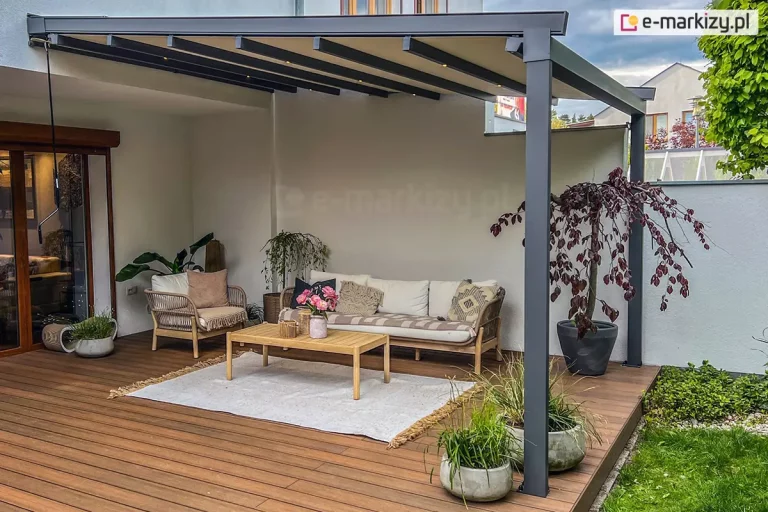 Zdjęcie prezentuje nowoczesną pergolę tarasową Aura o konstrukcji aluminiowej w kolorze ciemnego antracytu, zamontowaną do jasnej elewacji budynku. Pergola posiada zadaszenie tkaninowe w odcieniu beżowym, które jest rozpięte na poprzecznych profilach przeciwwietrznych. Charakterystycznym elementem tego modelu jest zintegrowane oświetlenie LED w formie punktowych diod umieszczonych w profilach podtrzymujących materiał, co tworzy nastrojową atmosferę po zmroku. Konstrukcja wspiera się na solidnych pionowych słupach o kwadratowym przekroju, usytuowanych przy zewnętrznej krawędzi tarasu. Po lewej stronie, przy ścianie budynku, widoczna jest długa korba służąca do manualnego sterowania mechanizmem składania dachu. Przestrzeń pod pergolą została zaaranżowana jako przytulny salon ogrodowy na drewnianym tarasie wykonanym z desek w ciepłym, brązowym kolorze. W centrum znajduje się niska sofa oraz fotel o konstrukcji z jasnego drewna i plecionki, wyłożone kremowymi poduszkami. Na sofie leżą dodatkowe poduszki dekoracyjne w odcieniach beżu i szarości, w tym jedna z etnicznym wzorem. Przed meblami rozłożono jasny, płasko tkany dywan z frędzlami, na którym stoi prostokątny, drewniany stolik kawowy ozdobiony wazonem z różowymi kwiatami. Wystrój tarasu uzupełniają liczne rośliny w donicach. Po prawej stronie, w dużej antracytowej donicy, rośnie ozdobne drzewko o ciemnobordowych liściach, prawdopodobnie buk pospolity w odmianie purpurowej. Wzdłuż krawędzi tarasu ustawiono kilka betonowych donic z ozdobnymi trawami i niską roślinnością. Tło stanowi biała ściana budynku oraz fragment ogrodu z zielonym trawnikiem i gęstymi krzewami widocznymi w oddali. Całość kompozycji łączy nowoczesny design systemów osłonowych z naturalnymi materiałami wykończeniowymi.