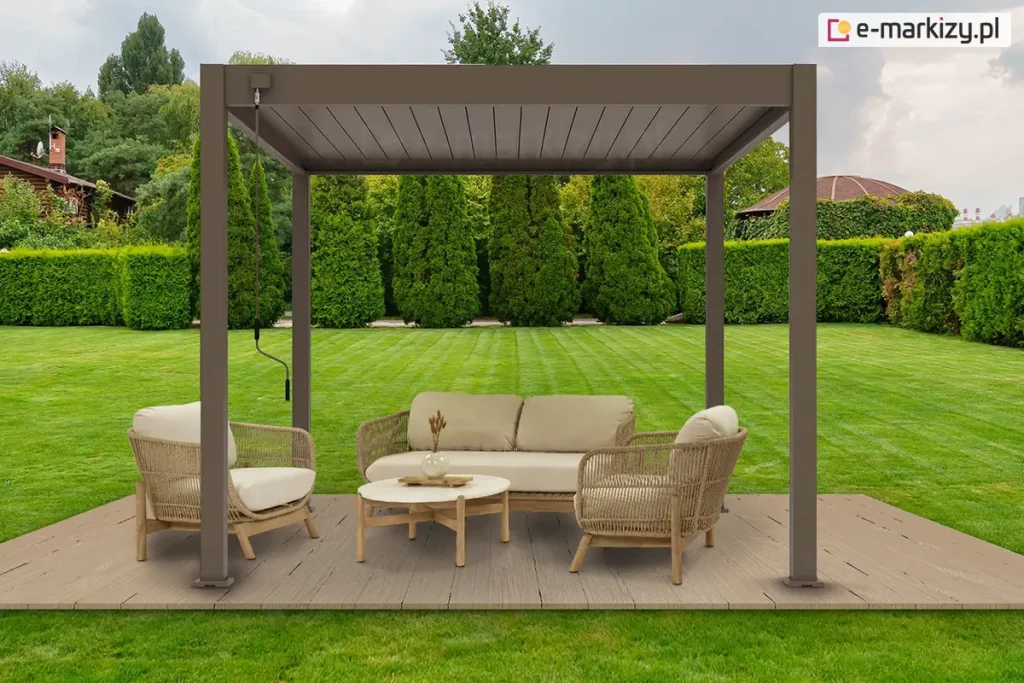 Podest z desek ułożony na trawniku w zadbanym ogrodzie z wysokimi tujami oraz przyciętym żywopłotem. Na podeście ustawiona pergola lamelowa Marlo w kolorze taupe z napędem ręcznym, za pomocą korby przymocowanej do konstrukcji. Pod szczelnie zamkniętym dachem pergoli Marlo ustawiono na podeście dwa fotele oraz dwuosobową kanapę z plecionym oparciem i drewnianymi nożkami. Pomiędzy nimi ustawiono niski stolik dopasowany stylem do siedzisk, na którym stoi wazon z ozdobna roślinką. Siedziska i oparcia foteli i sofy wyłożono miękkimi beżowymi poduszkami.