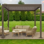 Podest z desek ułożony na trawniku w zadbanym ogrodzie z wysokimi tujami oraz przyciętym żywopłotem. Na podeście ustawiona pergola lamelowa Marlo w kolorze taupe z napędem ręcznym, za pomocą korby przymocowanej do konstrukcji. Pod szczelnie zamkniętym dachem pergoli Marlo ustawiono na podeście dwa fotele oraz dwuosobową kanapę z plecionym oparciem i drewnianymi nożkami. Pomiędzy nimi ustawiono niski stolik dopasowany stylem do siedzisk, na którym stoi wazon z ozdobna roślinką. Siedziska i oparcia foteli i sofy wyłożono miękkimi beżowymi poduszkami.