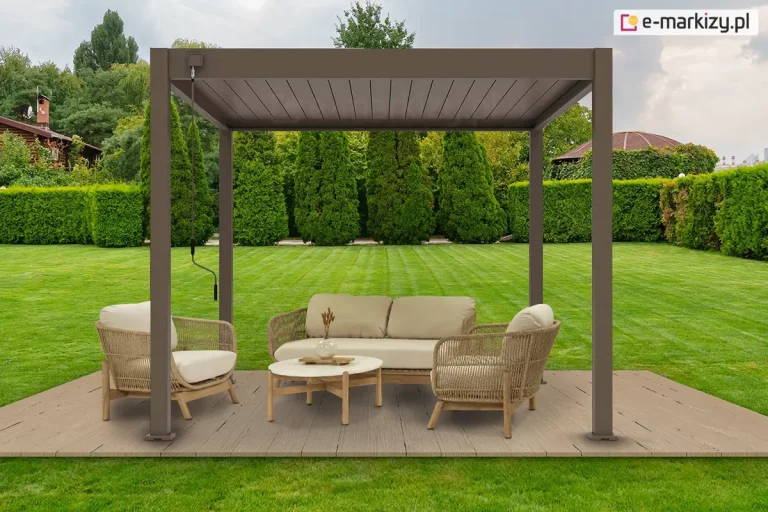 Podest z desek ułożony na trawniku w zadbanym ogrodzie z wysokimi tujami oraz przyciętym żywopłotem. Na podeście ustawiona pergola lamelowa Marlo w kolorze taupe z napędem ręcznym, za pomocą korby przymocowanej do konstrukcji. Pod szczelnie zamkniętym dachem pergoli Marlo ustawiono na podeście dwa fotele oraz dwuosobową kanapę z plecionym oparciem i drewnianymi nożkami. Pomiędzy nimi ustawiono niski stolik dopasowany stylem do siedzisk, na którym stoi wazon z ozdobna roślinką. Siedziska i oparcia foteli i sofy wyłożono miękkimi beżowymi poduszkami.