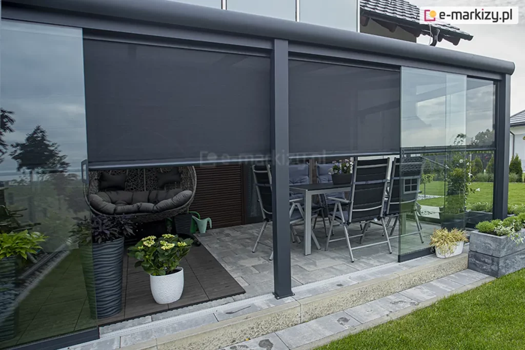 Zabudowany taras ogrodowy z przeszkloną konstrukcją aluminiową, wyposażony w rolety screenowe Refleksol 103 Selt w kolorze grafitowym, częściowo opuszczone w celu zapewnienia ochrony przeciwsłonecznej. Wnętrze tarasu urządzone jest jak przytulna strefa wypoczynkowa — po lewej stronie znajduje się nowoczesny fotel wiszący typu kokon z ciemnoszarymi poduszkami, ustawiony na drewnianym podeście. W centralnej części stoi zestaw mebli ogrodowych: metalowy stół i sześć składanych krzeseł w kolorze antracytowym z ciemnymi poduszkami. Taras otaczają liczne donice z roślinami ozdobnymi: białe i czarne doniczki z hortensjami, paprociami i inną zielenią ustawione są wzdłuż ścian i w rogu przy szklanej ścianie. Refleksol 103 to system idealny do ogrodów zimowych i pergoli — zapewnia skuteczną ochronę przed promieniowaniem UV, redukuje nagrzewanie wnętrza i zwiększa prywatność bez ograniczania widoku na zewnątrz.