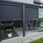 Zabudowany taras ogrodowy z przeszkloną konstrukcją aluminiową, wyposażony w rolety screenowe Refleksol 103 Selt w kolorze grafitowym, częściowo opuszczone w celu zapewnienia ochrony przeciwsłonecznej. Wnętrze tarasu urządzone jest jak przytulna strefa wypoczynkowa — po lewej stronie znajduje się nowoczesny fotel wiszący typu kokon z ciemnoszarymi poduszkami, ustawiony na drewnianym podeście. W centralnej części stoi zestaw mebli ogrodowych: metalowy stół i sześć składanych krzeseł w kolorze antracytowym z ciemnymi poduszkami. Taras otaczają liczne donice z roślinami ozdobnymi: białe i czarne doniczki z hortensjami, paprociami i inną zielenią ustawione są wzdłuż ścian i w rogu przy szklanej ścianie. Refleksol 103 to system idealny do ogrodów zimowych i pergoli — zapewnia skuteczną ochronę przed promieniowaniem UV, redukuje nagrzewanie wnętrza i zwiększa prywatność bez ograniczania widoku na zewnątrz.