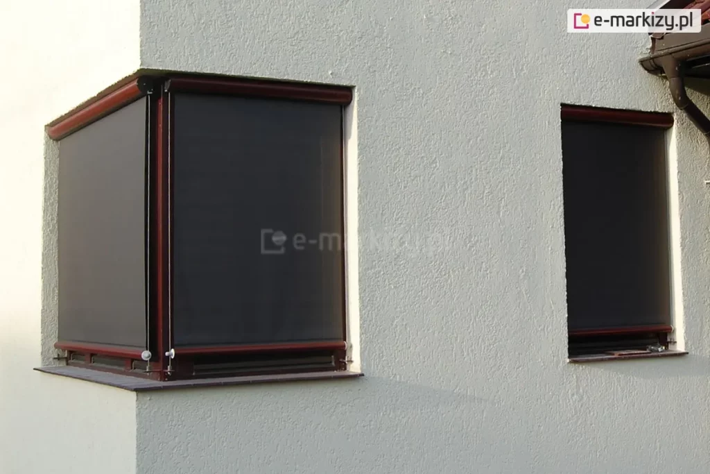 Narożnik budynku o białej fasadzie z widocznymi dwoma oknami: narożnym oraz wąskim okienkiem na prawej ścianie. Oba okna w kolorze brązowym osłonięto refleksolami 125 Selt w kolorystyce konstrukcji i tkaniny zbliżonej do barwy okien. Wszystkie okna są w pełni zasłonięte materiałem rolety screen.