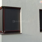 Narożnik budynku o białej fasadzie z widocznymi dwoma oknami: narożnym oraz wąskim okienkiem na prawej ścianie. Oba okna w kolorze brązowym osłonięto refleksolami 125 Selt w kolorystyce konstrukcji i tkaniny zbliżonej do barwy okien. Wszystkie okna są w pełni zasłonięte materiałem rolety screen.