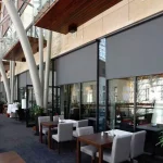 Wnętrze przeszklonego dziedzińca lub galerii handlowej, z widokiem na elegancką restaurację wkomponowaną w nowoczesną architekturę. Na dużych przeszkleniach zamontowane są zewnętrzne osłony przeciwsłoneczne Refleksol 90 Selt z tkaniną w kolorze ciemnoszarym, częściowo rozwinięte i osłaniające wnętrze lokalu. Rolety te mają formę screenów montowanych w kasetach, z pionowym prowadzeniem po bocznych prowadnicach. Po lewej i peawej stronie widoczne są stoliki z drewnianym blatem, tapicerowane krzesła w kolorze szarym oraz rośliny w donicach. Przestrzeń nad witrynami zdobią nowoczesne, łukowate podpory konstrukcyjne.