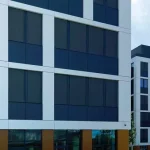 Widok na nowoczesną fasadę wielokondygnacyjnego budynku biurowego, wykończoną w geometrycznym układzie z naprzemiennie rozmieszczonymi polami w kolorze bieli i grafitu. Na każdym piętrze widoczne są zespolone pionowo okna, wyposażone w zewnętrzne osłony przeciwsłoneczne Refleksol 90 Selt. Rolety w ciemnym odcieniu szarości lub antracytu są całkowicie rozwinięte, tworząc jednolitą, nowoczesną powierzchnię zasłaniającą szyby. Budynek w tle utrzymany jest w tej samej stylistyce, co świadczy o spójnym projekcie architektonicznym. Dolna kondygnacja z dużymi przeszkleniami sugeruje przestrzeń komercyjną.