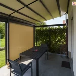 widok od wewnątrz na taras osłonięty pergolą Solid z dachem tkaninowym oraz rozwiniętą przednią roletą typu Refleksol. Konstrukcja pergoli w kolorze antracytowym wspiera beżowe, lekko nachylone poszycie dachowe, które chroni strefę wypoczynkową przed słońcem. W świetle frontowego przejścia znajduje się pionowa roleta w kolorze piaskowym, która częściowo przysłania widok na ogród, zapewniając ochronę przed promieniami słonecznymi i wiatrem. Pod zadaszeniem ustawiono prostokątny, czarny stół z dopasowanymi nowoczesnymi krzesłami ogrodowymi na tle szarej, wielkoformatowej kostki tarasowej. W tle, za pergolą, widoczny jest zielony trawnik, ogrodzenie panelowe oraz drewniana kratka z gęstym pnączem.