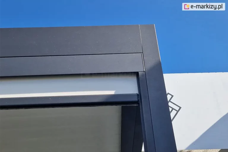 Zbliżenie techniczne na górny narożnik zewnętrznego refleksola Ziiip 120 Selt w kolorze antracytowym zamocowanego do pergoli w tym samym kolorze. Zdjęcie wykonane pod kątem na tle czystego, błękitnego nieba, prezentuje precyzyjne łączenie elementów aluminiowej obudowy. Widoczna jest kwadratowa kaseta górna z wyraźną linią podziału między pokrywą a korpusem, połączona z pionową prowadnicą boczną. Z wnętrza kasety wysunięta jest pozioma listwa dolna (obciążnik) w tym samym kolorze co konstrukcja, pod którą znajduje się fragment jasnej, technicznej tkaniny osłonowej. Całość konstrukcji posiada matowe wykończenie lakiernicze o delikatnej strukturze, charakterystyczne dla nowoczesnej stolarki aluminiowej.