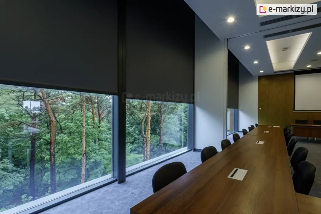 Widok wnętrza eleganckiej sali konferencyjnej z wysokimi przeszkleniami, na których zastosowano zewnętrzne rolety przeciwsłoneczne Refleksol 103 Selt. Czarna tkanina screenowa częściowo opuszczona, zapewnia skuteczną ochronę przed światłem i nagrzewaniem, jednocześnie umożliwiając podgląd na zielony krajobraz za oknem. Pomieszczenie wyróżnia się stonowaną kolorystyką, nowoczesnym oświetleniem sufitowym i dużym, drewnianym stołem – idealnym do pracy zespołowej. System refleksol idealnie sprawdza się w przestrzeniach komercyjnych, gdzie estetyka musi iść w parze z funkcjonalnością.