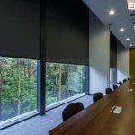Widok wnętrza eleganckiej sali konferencyjnej z wysokimi przeszkleniami, na których zastosowano zewnętrzne rolety przeciwsłoneczne Refleksol 103 Selt. Czarna tkanina screenowa częściowo opuszczona, zapewnia skuteczną ochronę przed światłem i nagrzewaniem, jednocześnie umożliwiając podgląd na zielony krajobraz za oknem. Pomieszczenie wyróżnia się stonowaną kolorystyką, nowoczesnym oświetleniem sufitowym i dużym, drewnianym stołem – idealnym do pracy zespołowej. System refleksol idealnie sprawdza się w przestrzeniach komercyjnych, gdzie estetyka musi iść w parze z funkcjonalnością.