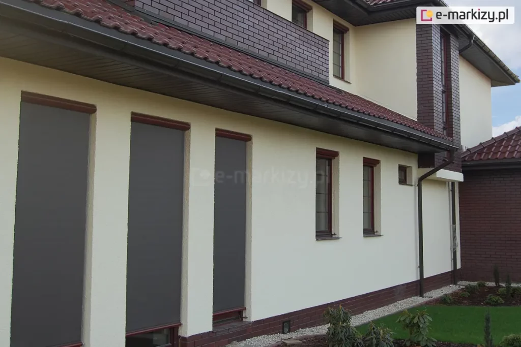 Boczną elewacja domu jednorodzinnego z trzema dużymi oknami zabezpieczonymi zewnętrznymi osłonami typu Refleksol 76. Rolety screenowe zostały całkowicie opuszczone i wykonane są z półprzezroczystej tkaniny w kolorze szarym oraz posiadają prowadnice linkowe mocowane do ramy okna. U góry widoczne są smukłe kasety w kolorze ciemnobrązowym z połyskiem, dopasowanym do ram okiennych. Ściana wykończona jest jasnym, kremowym tynkiem, a cokół oraz elementy narożne wykonane są z czerwono-brązowej cegły klinkierowej. Budynek posiada dach z dachówką ceramiczną w odcieniu bordo oraz klasyczne rynny i obróbki blacharskie. Tło stanowi zadbany ogród z niskimi roślinami ozdobnymi i trawnikiem. Refleksol 76 to nowoczesny system rolet screenowych, montowanych na elewacji, który skutecznie chroni wnętrze przed przegrzewaniem, jednocześnie nie blokując całkowicie dostępu światła dziennego.