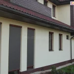 Boczną elewacja domu jednorodzinnego z trzema dużymi oknami zabezpieczonymi zewnętrznymi osłonami typu Refleksol 76. Rolety screenowe zostały całkowicie opuszczone i wykonane są z półprzezroczystej tkaniny w kolorze szarym oraz posiadają prowadnice linkowe mocowane do ramy okna. U góry widoczne są smukłe kasety w kolorze ciemnobrązowym z połyskiem, dopasowanym do ram okiennych. Ściana wykończona jest jasnym, kremowym tynkiem, a cokół oraz elementy narożne wykonane są z czerwono-brązowej cegły klinkierowej. Budynek posiada dach z dachówką ceramiczną w odcieniu bordo oraz klasyczne rynny i obróbki blacharskie. Tło stanowi zadbany ogród z niskimi roślinami ozdobnymi i trawnikiem. Refleksol 76 to nowoczesny system rolet screenowych, montowanych na elewacji, który skutecznie chroni wnętrze przed przegrzewaniem, jednocześnie nie blokując całkowicie dostępu światła dziennego.