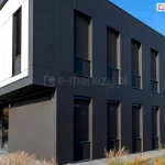 Nowoczesny budynek biurowy z zamontowanymi refleksolami w dolnej części elewacji od frontu oraz na prawej ścianie budynku. Rolety zewnętrzne w ciemnym kolorze osłaniają okna, zapewniając prywatność i ochronę przed słońcem, jednocześnie umożliwiając widoczność na zewnątrz.