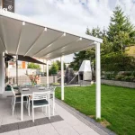 Biała pergola tarasowa Tarasola Simple o konstrukcji przyściennej zamontowana nad nowoczesnym tarasem wyłożonym jasnymi płytami chodnikowymi z ciemniejszymi pasami dekoracyjnymi. Zadaszenie pergoli wykonane jest z trójwarstwowej tkaniny technicznej ułożonej w charakterystyczne, równolegle biegnące fałdy, które są wsparte na poprzecznych profilach aluminiowych. Konstrukcja opiera się z jednej strony na elewacji budynku wykończonej panelami o strukturze drewna, a z drugiej strony na dwóch pionowych, kwadratowych słupach nośnych w kolorze białym. Pod pergolą znajduje się zestaw mebli ogrodowych składający się z dużego szarego stołu oraz sześciu białych krzeseł z ciemnymi poduszkami na siedziskach. Na stole ustawiony jest wazon z kwiatami oraz zastawa obiadowa. W tle widoczny jest zadbany trawnik, murowany grill ogrodowy o jasnej kolorystyce oraz niska roślinność i drzewa iglaste oddzielające posesję od sąsiednich budynków. Całość kompozycji przedstawiona jest w pełnym świetle dziennym przy lekko zachmurzonym niebie.