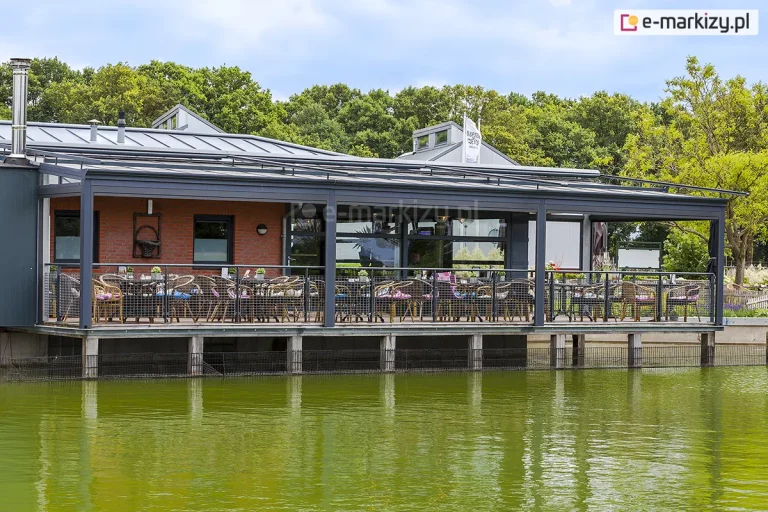 szerokie, modułowe zadaszenie tarasowe model Terano zainstalowane nad zewnętrznym podestem restauracji zlokalizowanej bezpośrednio nad zbiornikiem wodnym. Konstrukcja o znacznej rozpiętości wykonana jest z profili aluminiowych w kolorze antracytowym i składa się z kilku sekcji wspartych na szeregu pionowych słupów nośnych. Każda sekcja dachu posiada wypełnienie z przezroczystych tafli szklanych. Nad szklanym dachem zamontowano zewnętrzny system osłon przeciwsłonecznych w postaci rolet rzymskich na podwyższonych prowadnicach, co umożliwia regulację nasłonecznienia tarasu. Podest restauracji jest wyniesiony nad lustro wody na betonowych i metalowych palach, a jego obwód zabezpieczono balustradą z metalowej siatki rozpiętej w czarnych ramach. W strefie jadalnianej ustawiono liczne stoliki z plecionymi krzesłami ogrodowymi, wyposażonymi w kolorowe poduszki. Budynek restauracji posiada elewację z czerwonej cegły oraz duże przeszklenia drzwi i okien. W tle widoczna jest gęsta linia drzew oraz pochmurne niebo, które odbija się w zielonkawej wodzie.