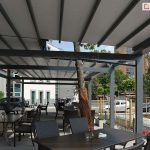 Wnętrze rozbudowanej, wielomodułowej pergoli tarasowej Solid Selt o konstrukcji aluminiowej w kolorze antracytowym, zaadaptowanej na ogródek restauracyjny. Zadaszenie składa się z rozsuwanego, jasnoszarego materiału zamontowanego na prowadnicach. Pod pergolą ustawiono liczne ciemne stoły z dopasowanymi fotelami z technorattanu. Konstrukcja została zaprojektowana w sposób omijający rosnące drzewo, którego pień zabezpieczono brązową matą osłonową. W tle widoczna jest nowoczesna architektura miejska: biały budynek biurowy, parking z samochodami oraz młode drzewka w ozdobnych palisadach. Po prawej stronie w oddali majaczy ceglana wieża o stożkowym dachu pod błękitnym niebem. Na stolikach restauracyjnych widoczne są drobne dekoracje, takie jak małe wazony z kwiatami.