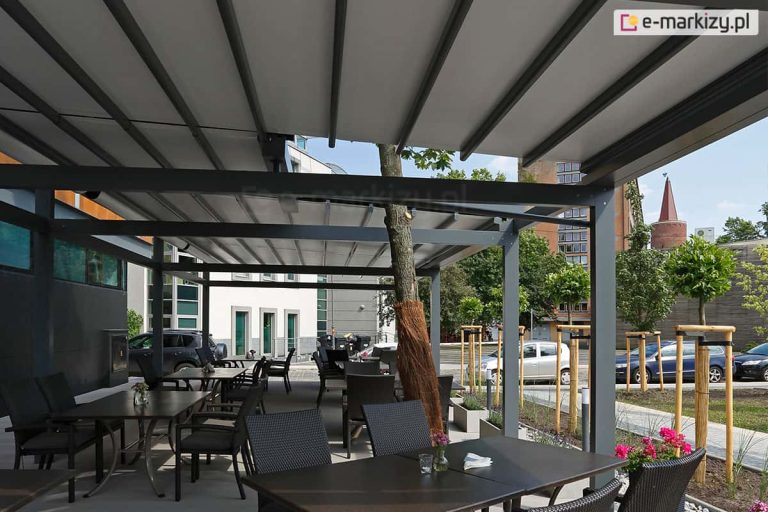 Wnętrze rozbudowanej, wielomodułowej pergoli tarasowej Solid Selt o konstrukcji aluminiowej w kolorze antracytowym, zaadaptowanej na ogródek restauracyjny. Zadaszenie składa się z rozsuwanego, jasnoszarego materiału zamontowanego na prowadnicach. Pod pergolą ustawiono liczne ciemne stoły z dopasowanymi fotelami z technorattanu. Konstrukcja została zaprojektowana w sposób omijający rosnące drzewo, którego pień zabezpieczono brązową matą osłonową. W tle widoczna jest nowoczesna architektura miejska: biały budynek biurowy, parking z samochodami oraz młode drzewka w ozdobnych palisadach. Po prawej stronie w oddali majaczy ceglana wieża o stożkowym dachu pod błękitnym niebem. Na stolikach restauracyjnych widoczne są drobne dekoracje, takie jak małe wazony z kwiatami.