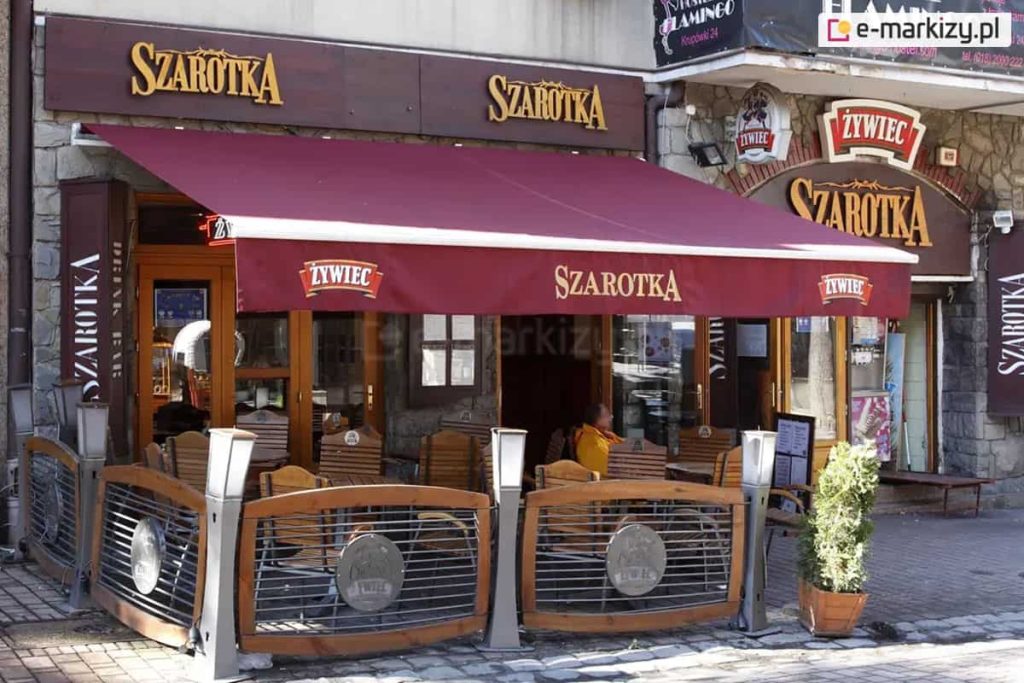 Markiza tarasowa Australia w kolorze białym z poszyciem w kolorze brodowo-czerwonym oraz z nadrukiem na przedłużonej falbanie o treści "szarotka" oraz umieszczonymi na skrajach logo "Żywiec" rozwinięta nad stolikami kawiarni szarotka, które wystawione zostały przed wejściem do lokalu i otoczone płotkami z reklamą połączonymi słupkami w postaci lamp ogrodowych.