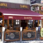 Markiza tarasowa Australia w kolorze białym z poszyciem w kolorze brodowo-czerwonym oraz z nadrukiem na przedłużonej falbanie o treści "szarotka" oraz umieszczonymi na skrajach logo "Żywiec" rozwinięta nad stolikami kawiarni szarotka, które wystawione zostały przed wejściem do lokalu i otoczone płotkami z reklamą połączonymi słupkami w postaci lamp ogrodowych.