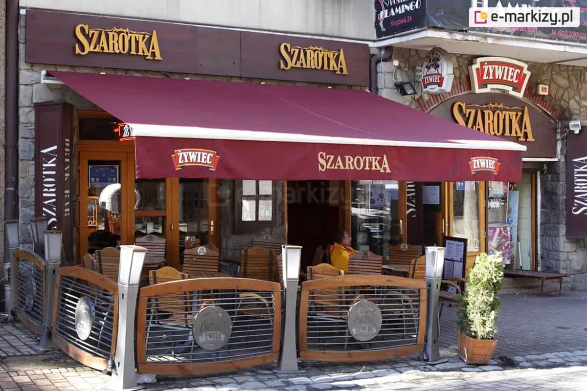 Markiza tarasowa Australia w kolorze białym z poszyciem w kolorze brodowo-czerwonym oraz z nadrukiem na przedłużonej falbanie o treści "szarotka" oraz umieszczonymi na skrajach logo "Żywiec" rozwinięta nad stolikami kawiarni szarotka, które wystawione zostały przed wejściem do lokalu i otoczone płotkami z reklamą połączonymi słupkami w postaci lamp ogrodowych.