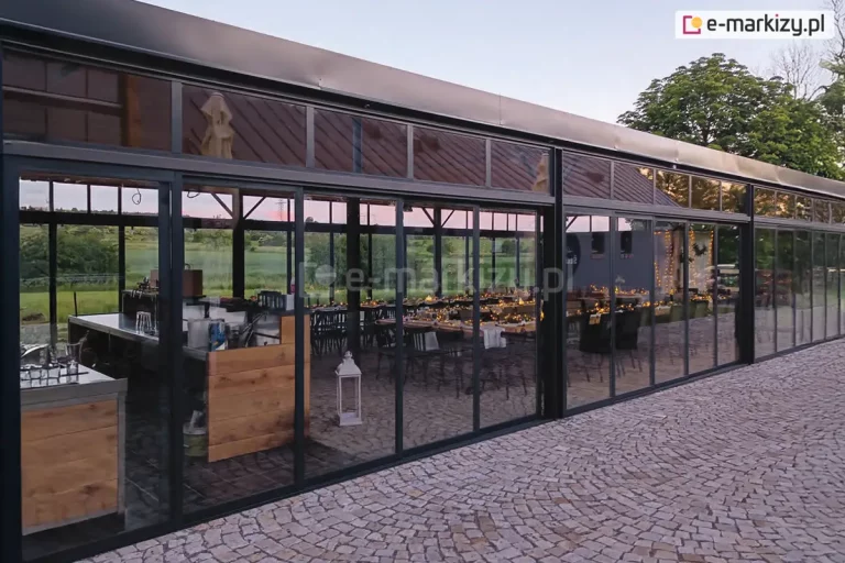 Zewnętrzna perspektywa nowoczesnej, wielkogabarytowej zabudowy szklanej restauracji wykonanej w systemie przesuwnym Sotro o ciemnej, grafitowej konstrukcji aluminiowej. Zdjęcie prezentuje długą linię przeszklonych segmentów, które obejmują zarówno dolne panele ruchome, jak i górne naświetla stałe z lekko przyciemnianym szkłem refleksyjnym, w którym odbija się dach sąsiedniego budynku. Przez transparentne tafle dolnych skrzydeł widoczne jest w pełni wyposażone wnętrze z barem wykonanym z drewna i stali nierdzewnej oraz długie stoły przygotowane do uroczystości, ozdobione dekoracyjnym oświetleniem. Całość konstrukcji osadzona jest na nawierzchni z jasnej kostki granitowej ułożonej w tradycyjny wzór. System charakteryzuje się dużymi przeszkleniami i wąskimi profilami, co nadaje bryle budynku lekkości i pozwala na całkowite otwarcie przestrzeni w sezonie letnim.