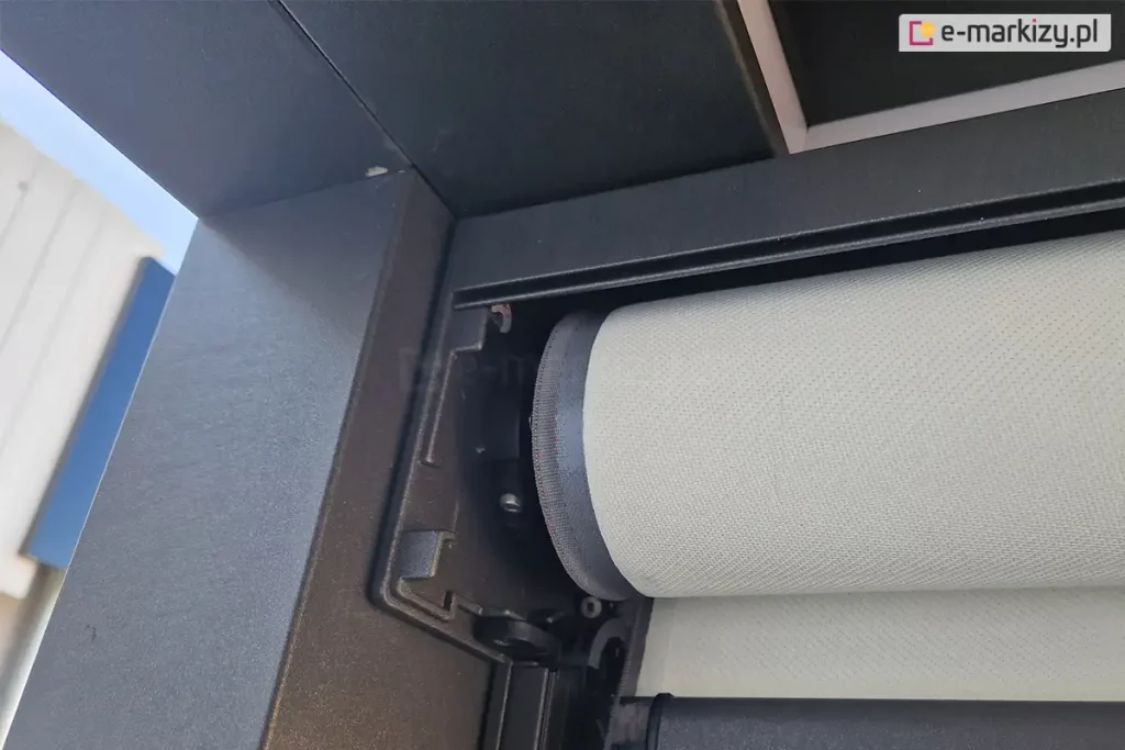 Zbliżenie techniczne na otwartą kasetę zewnętrznej rolety materiałowej typu screen, prezentujące mechanizm nawojowy. Na zdjęciu widoczna jest rura nawojowa, na którą nawinięta jest jasna tkanina techniczna o wyraźnej, perforowanej strukturze. Materiał wykończony jest na krawędzi szarym wzmocnieniem - Ziiip. Konstrukcja kasety oraz boczny uchwyt mocujący wał wykonane są z aluminium lakierowanego proszkowo na kolor ciemnoszary (antracyt). Widoczne są detale montażowe, w tym gniazdo osadzenia rury oraz śruby imbusowe mocujące elementy mechaniczne. Zdjęcie wykonane z perspektywy dolnej, ukazujące precyzyjne dopasowanie komponentów systemu osłonowego. Tło stanowi fragment elewacji budynku oraz niebo, co podkreśla natynkowy charakter montażu produktu.