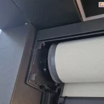 Zbliżenie techniczne na otwartą kasetę zewnętrznej rolety materiałowej typu screen, prezentujące mechanizm nawojowy. Na zdjęciu widoczna jest rura nawojowa, na którą nawinięta jest jasna tkanina techniczna o wyraźnej, perforowanej strukturze. Materiał wykończony jest na krawędzi szarym wzmocnieniem - Ziiip. Konstrukcja kasety oraz boczny uchwyt mocujący wał wykonane są z aluminium lakierowanego proszkowo na kolor ciemnoszary (antracyt). Widoczne są detale montażowe, w tym gniazdo osadzenia rury oraz śruby imbusowe mocujące elementy mechaniczne. Zdjęcie wykonane z perspektywy dolnej, ukazujące precyzyjne dopasowanie komponentów systemu osłonowego. Tło stanowi fragment elewacji budynku oraz niebo, co podkreśla natynkowy charakter montażu produktu.