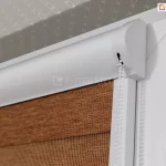 Zbliżenie na prawy górny narożnik białego okna PCV z zamontowaną roletą dzień i noc w systemie kasetowym. Zdjęcie prezentuje profil białej kasety osłonowej o ergonomicznym, zaokrąglonym kształcie, która jest zamontowana nawierzchniowo do ramy skrzydła okiennego. Z boku kasety, przez specjalny pionowy otwór w boczku, wyprowadzony jest biały, plastikowy łańcuszek koralikowy służący do płynnej regulacji wysokości rolety. Poniżej kasety znajduje się pionowa biała prowadnica przestrzenna, która utrzymuje materiał blisko szyby. Tkanina rolety układa się w poziome pasy: widoczny jest fragment szerokiego, nieprzeziernego pasa w kolorze ceglanego, melanżowego brązu oraz pas transparentnej, drobnej siateczki. Po prawej stronie kadru widoczny jest fragment ramy okiennej z zawiasem. W górnej części zdjęcia, ponad oknem, zarysowuje się wnęka okienna wykończona tapetą o subtelnym, szarym wzorze geometrycznym. Oświetlenie podkreśla gładką powierzchnię osprzętu z tworzywa oraz wyraźną fakturę splotu tkaniny.