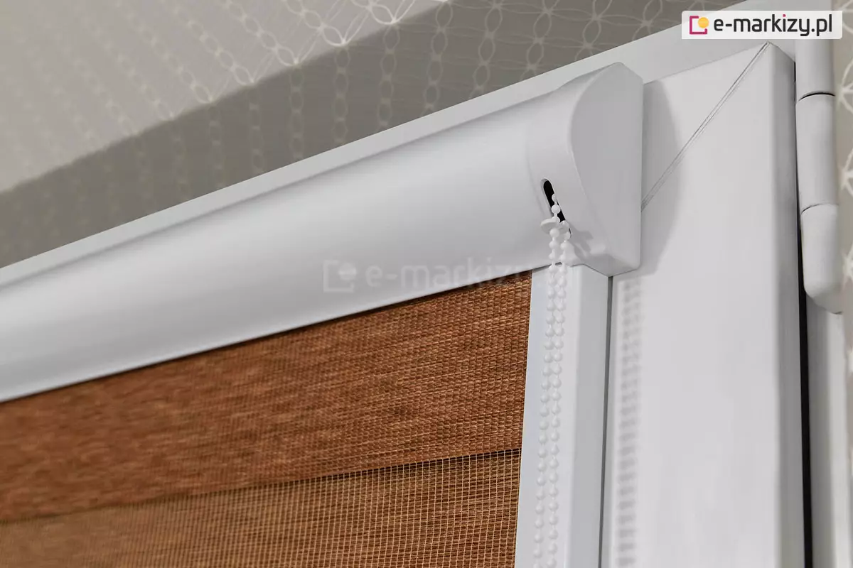 Zbliżenie na prawy górny narożnik białego okna PCV z zamontowaną roletą dzień i noc w systemie kasetowym. Zdjęcie prezentuje profil białej kasety osłonowej o ergonomicznym, zaokrąglonym kształcie, która jest zamontowana nawierzchniowo do ramy skrzydła okiennego. Z boku kasety, przez specjalny pionowy otwór w boczku, wyprowadzony jest biały, plastikowy łańcuszek koralikowy służący do płynnej regulacji wysokości rolety. Poniżej kasety znajduje się pionowa biała prowadnica przestrzenna, która utrzymuje materiał blisko szyby. Tkanina rolety układa się w poziome pasy: widoczny jest fragment szerokiego, nieprzeziernego pasa w kolorze ceglanego, melanżowego brązu oraz pas transparentnej, drobnej siateczki. Po prawej stronie kadru widoczny jest fragment ramy okiennej z zawiasem. W górnej części zdjęcia, ponad oknem, zarysowuje się wnęka okienna wykończona tapetą o subtelnym, szarym wzorze geometrycznym. Oświetlenie podkreśla gładką powierzchnię osprzętu z tworzywa oraz wyraźną fakturę splotu tkaniny.