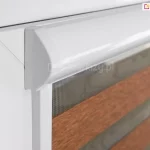 Zbliżenie na lewy górny narożnik białego okna PCV, na którym zamontowano system rolety dzień i noc w pełnej kasecie. Fotografia pod kątem prezentuje profil białej, aluminiowej kasety o wyraźnie zaokrąglonym froncie i płaskim boku, która ściśle przylega do ramy okiennej. Bezpośrednio pod kasetą widoczna jest pionowa, biała prowadnica przestrzenna, w której osadzony jest materiał rolety. Tkanina składa się z poziomych, naprzemiennych pasów: górny pas to transparentna, drobna siateczka w kolorze szarym, pod którą znajduje się szeroki, nieprzezierny pas materiału w kolorze ceglanego brązu o melanżowej strukturze. W tle, za transparentną częścią rolety, widoczny jest fragment ramy okiennej i delikatny zarys szyby. Zdjęcie wykonane w jasnym pomieszczeniu, z dużą dbałością o detale techniczne łączenia kasety z prowadnicą oraz teksturę zastosowanych materiałów.