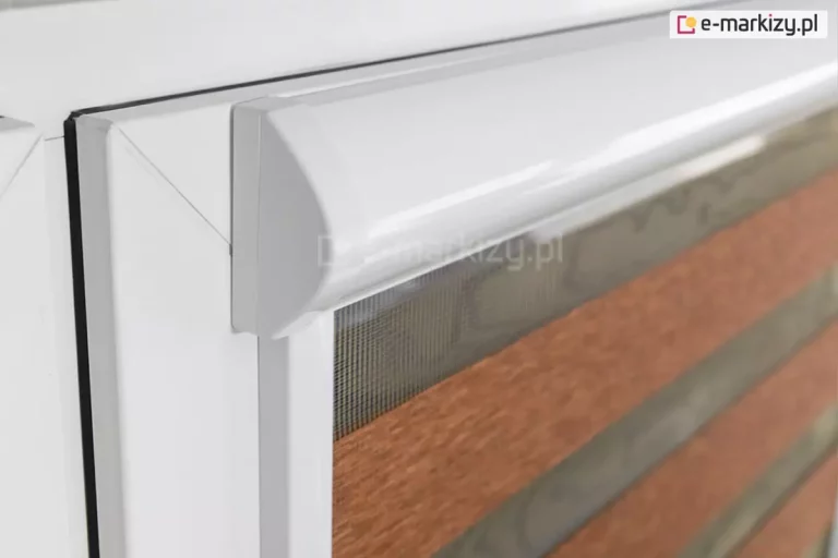 Zbliżenie na lewy górny narożnik białego okna PCV, na którym zamontowano system rolety dzień i noc w pełnej kasecie. Fotografia pod kątem prezentuje profil białej, aluminiowej kasety o wyraźnie zaokrąglonym froncie i płaskim boku, która ściśle przylega do ramy okiennej. Bezpośrednio pod kasetą widoczna jest pionowa, biała prowadnica przestrzenna, w której osadzony jest materiał rolety. Tkanina składa się z poziomych, naprzemiennych pasów: górny pas to transparentna, drobna siateczka w kolorze szarym, pod którą znajduje się szeroki, nieprzezierny pas materiału w kolorze ceglanego brązu o melanżowej strukturze. W tle, za transparentną częścią rolety, widoczny jest fragment ramy okiennej i delikatny zarys szyby. Zdjęcie wykonane w jasnym pomieszczeniu, z dużą dbałością o detale techniczne łączenia kasety z prowadnicą oraz teksturę zastosowanych materiałów.