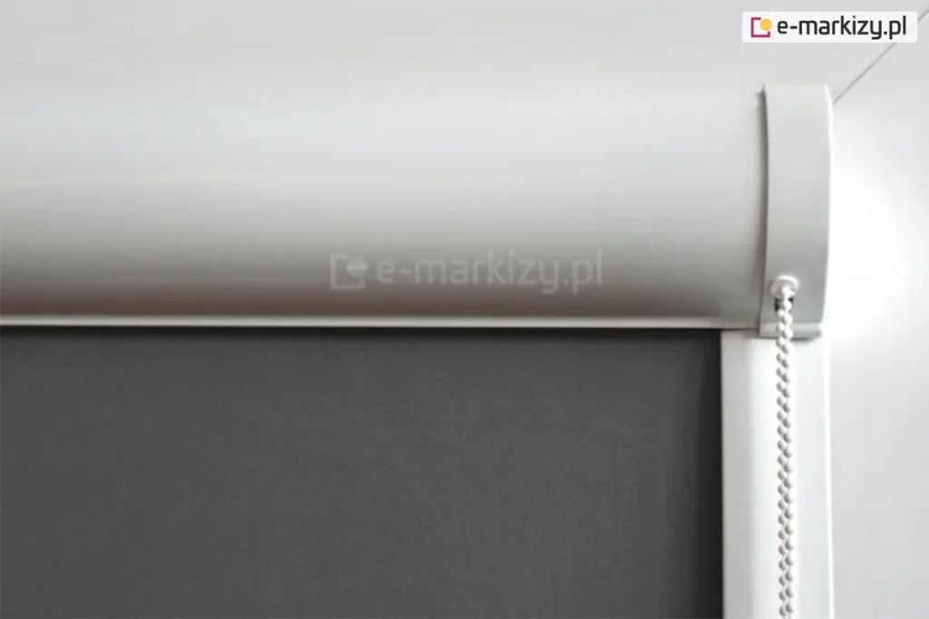 Zbliżenie na górny profil rolety wewnętrznej w białej, zaokrąglonej kasecie osłaniającej mechanizm nawojowy. Pod kasetą znajduje się gładka tkanina w kolorze ciemnoszarym lub antracytowym o gęstym, jednolitym splocie, który zapewnia maksymalne zaciemnienie pomieszczenia. Po prawej stronie widoczny jest biały łańcuszek koralikowy służący do sterowania wysokością rolety, który wychodzi z bocznego elementu wykończeniowego kasety. Pionowa, biała prowadnica boczna biegnie w dół wzdłuż krawędzi materiału, zapewniając jego stabilne przyleganie. Całość konstrukcji, obejmująca kasetę, boczki oraz prowadnice, jest wykonana z białego tworzywa lub lakierowanego aluminium o satynowym wykończeniu. Tło zdjęcia jest jasne i neutralne, co uwydatnia techniczne detale montażowe oraz kontrast między jasną obudową a ciemnym materiałem przesłonowym.